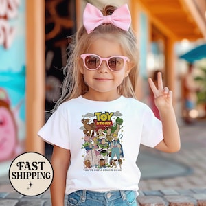 Puede incluir: Una ni&ntilde;a peque&ntilde;a con una camiseta blanca con un gr&aacute;fico colorido de "Toy Story". La camiseta presenta personajes de la pel&iacute;cula y el texto "You've Got a Friend in Me". Tambi&eacute;n lleva gafas de sol rosas y un lazo rosa.