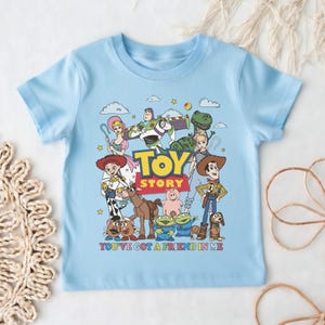 Op de afbeelding: Lichtblauw T-shirt met een kleurrijke afbeelding van personages uit de film "Toy Story". Het ontwerp bevat de tekst "TOY STORY" en "YOU'VE GOT A FRIEND IN ME."