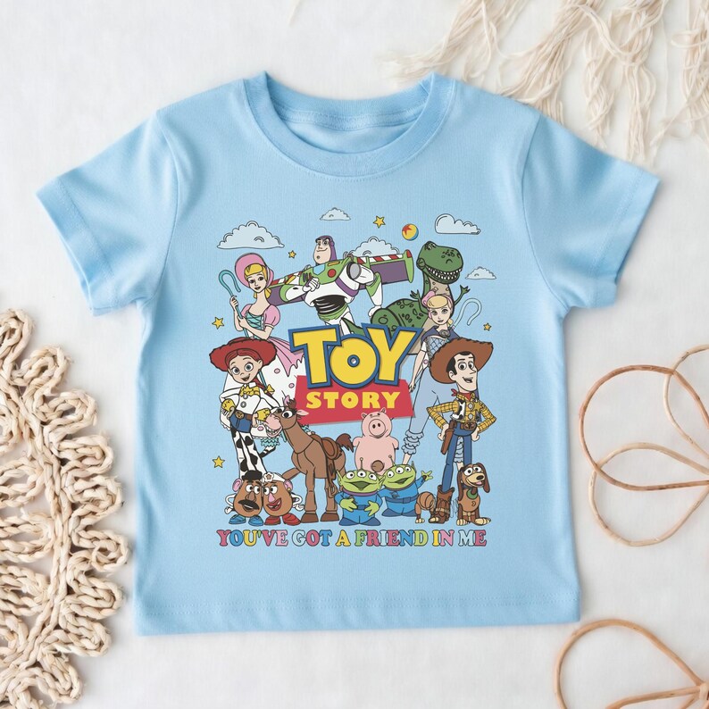 Op de afbeelding: Lichtblauw T-shirt met een kleurrijke afbeelding van personages uit de film "Toy Story". Het ontwerp bevat de tekst "TOY STORY" en "YOU'VE GOT A FRIEND IN ME."