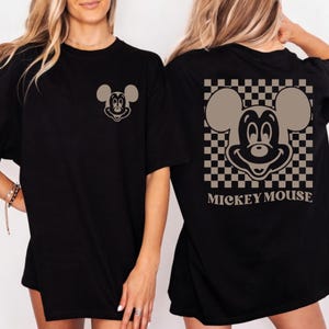 May include: Black t-shirt featuring a Mickey Mouse graphic. The front of the shirt has a small Mickey Mouse design, while the back showcases a larger Mickey Mouse image within a checkered pattern, with the words "Mickey Mouse" below.