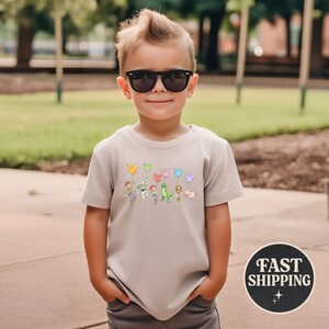 K&ouml;nnte beinhalten: Beigefarbenes T-Shirt mit einer Grafik von Zeichentrickfiguren, die Luftballons halten. Die Ballons sind in verschiedenen Farben und Formen gehalten. Das Shirt hat einen Rundhalsausschnitt und kurze &Auml;rmel. Das Bild enth&auml;lt auch eine "FAST SHIPPING" Grafik.