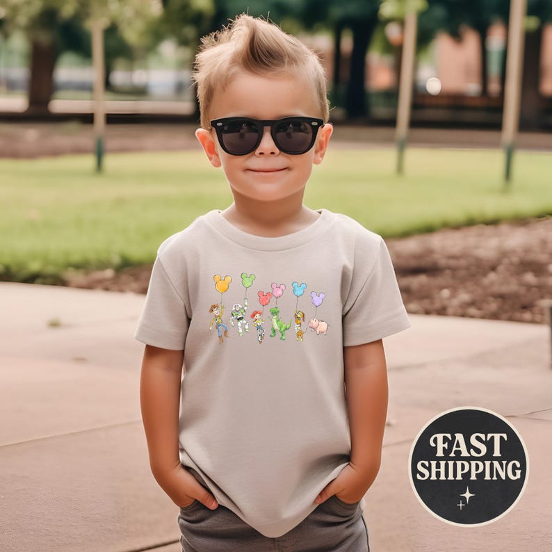 K&ouml;nnte beinhalten: Beigefarbenes T-Shirt mit einer Grafik von Zeichentrickfiguren, die Luftballons halten. Die Ballons sind in verschiedenen Farben und Formen gehalten. Das Shirt hat einen Rundhalsausschnitt und kurze &Auml;rmel. Das Bild enth&auml;lt auch eine "FAST SHIPPING" Grafik.