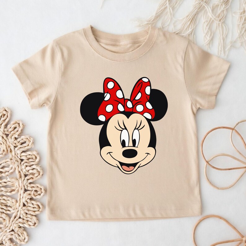 Może przedstawiać: Beżowa koszulka z grafiką Myszki Minnie. Projekt przedstawia twarz Minnie, w tym czerwoną kokardę w białe grochy, czarne uszy i uśmiechniętą twarz. Koszulka wykonana jest z miękkiej tkaniny w neutralnym kolorze.