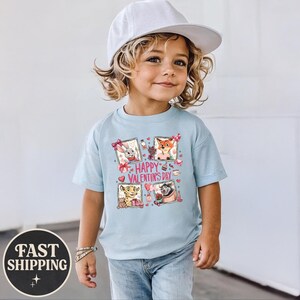 May include: A light blue t-shirt featuring four framed cartoon characters with Valentine's Day decorations. The shirt has the text "Happy Valentine's Day" in the center. The child is wearing a white cap and blue jeans.