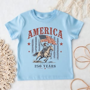 May include: A light blue short-sleeve t-shirt featuring a graphic print of a person riding a horse while holding an American flag. The text reads AMERICA, SINCE 1776, and 250 YEARS. The design includes red stars and a faded vertical striped background.