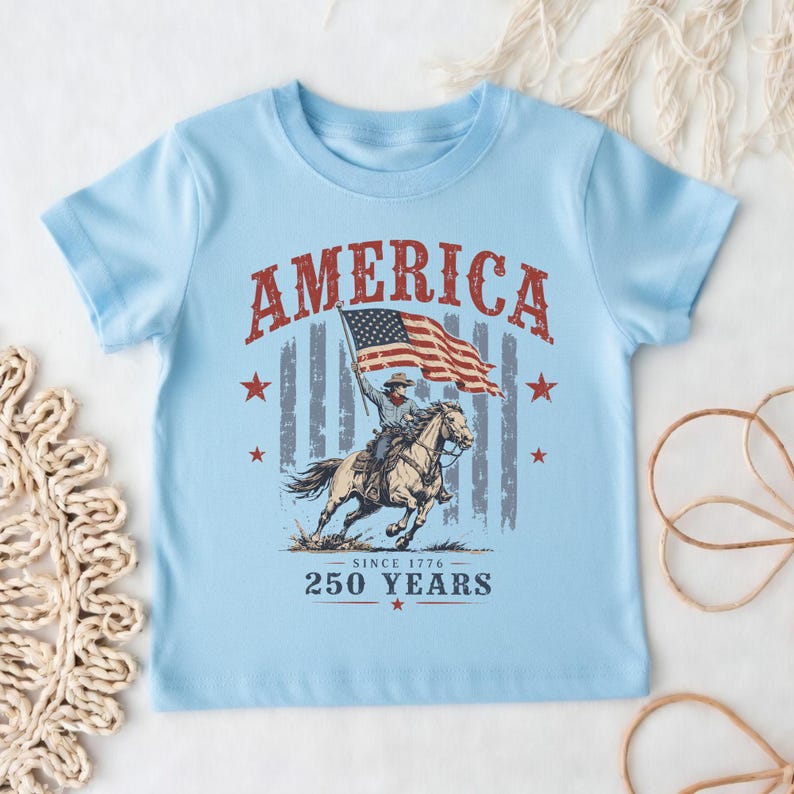 May include: A light blue short-sleeve t-shirt featuring a graphic print of a person riding a horse while holding an American flag. The text reads AMERICA, SINCE 1776, and 250 YEARS. The design includes red stars and a faded vertical striped background.