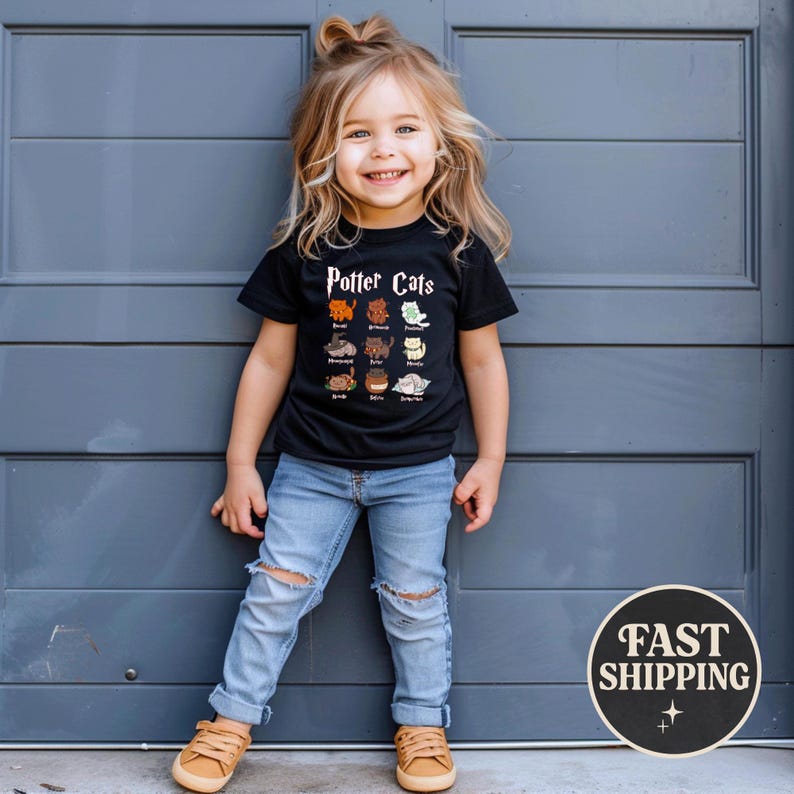 May include: A black t-shirt with the text "Potter Cats" and illustrations of various cats. The shirt is worn by a child with blonde hair and blue jeans. The child is smiling and standing in front of a gray door.