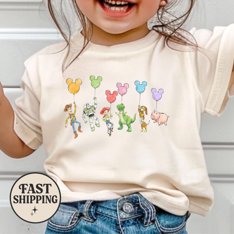 K&ouml;nnte beinhalten: Ein cremefarbenes T-Shirt mit einer bunten Grafik von Toy Story-Charakteren, die Mickey Mouse-f&ouml;rmige Ballons halten. Zu den Figuren geh&ouml;ren Woody, Buzz Lightyear, Jessie, Rex, Slinky Dog, Hamm und andere. Das Shirt hat einen Rundhalsausschnitt und kurze &Auml;rmel.