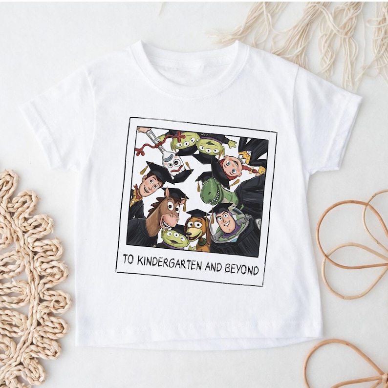 May include: A white short-sleeve t-shirt featuring a graphic print of a polaroid-style frame containing cartoon characters wearing graduation caps. The text below the characters reads TO KINDERGARTEN AND BEYOND in a black sans-serif font.