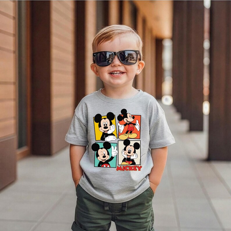 May include: A child wearing sunglasses and a grey t-shirt with four Mickey Mouse images. The word "Mickey" is printed in red. The child is also wearing green shorts. The background is a blurred outdoor setting.