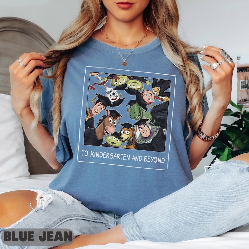 May include: A blue t-shirt featuring a square graphic of Toy Story characters wearing graduation caps. The characters include Woody, Buzz Lightyear, Rex, and others. Below the graphic, the text reads TO KINDERGARTEN AND BEYOND.