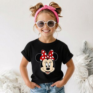 Może przedstawiać: Czarny t-shirt z motywem Myszki Minnie, z czerwoną kokardą w białe grochy. Dziecko ma na sobie niebieskie dżinsy, r&oacute;żową opaskę na głowę i białe okulary przeciwsłoneczne. Koszulka ma podwinięte rękawy.