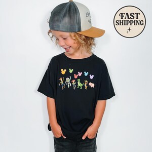 K&ouml;nnte beinhalten: Ein schwarzes T-Shirt mit bunten Zeichentrickfiguren und Mickey Mouse-f&ouml;rmigen Ballons. Das T-Shirt wird mit einer Trucker-M&uuml;tze mit braunem Schirm und grauem Mesh-R&uuml;cken getragen.