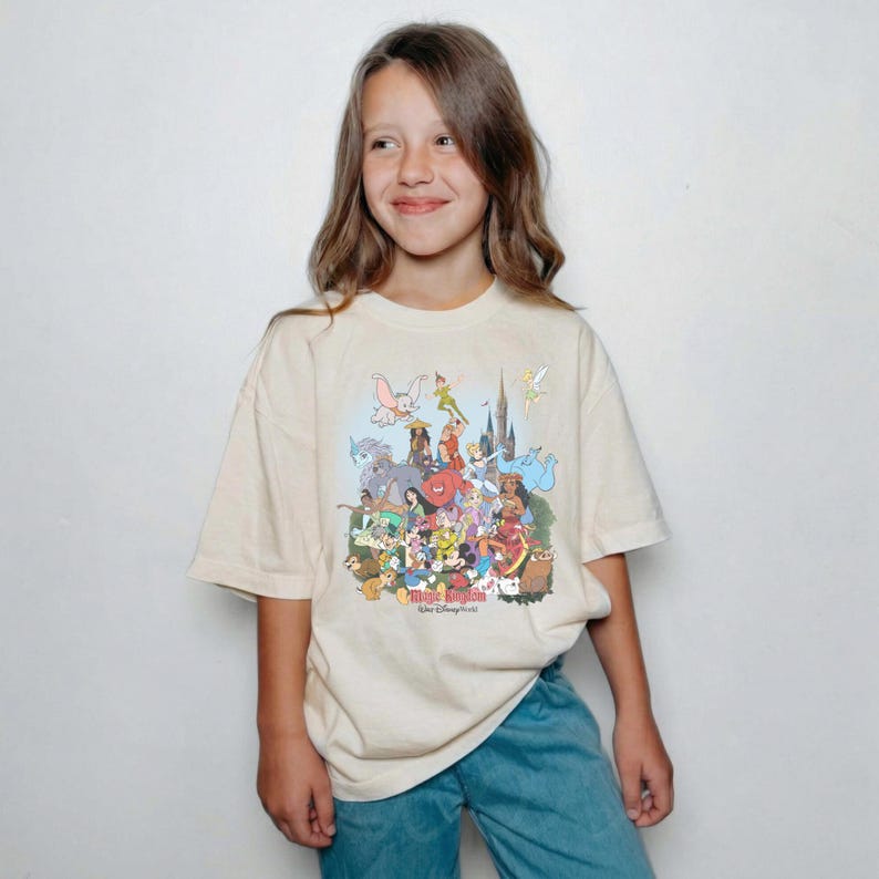 May include: Cream-colored t-shirt featuring a colorful graphic of Disney characters, including Mickey Mouse, Peter Pan, and Cinderella. The shirt has the words "Magic Kingdom" and "Walt Disney World" printed on it. The shirt is worn by a person.