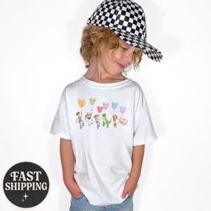 K&ouml;nnte beinhalten: Ein wei&szlig;es T-Shirt mit einer bunten Grafik von Toy Story-Charakteren, die Mickey Mouse-f&ouml;rmige Ballons halten. Das Kind tr&auml;gt eine schwarz-wei&szlig; karierte Baseballkappe. Das Shirt ist ein l&auml;ssiger Stil.
