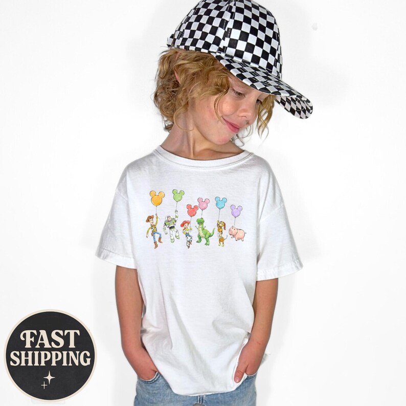 K&ouml;nnte beinhalten: Ein wei&szlig;es T-Shirt mit einer bunten Grafik von Toy Story-Charakteren, die Mickey Mouse-f&ouml;rmige Ballons halten. Das Kind tr&auml;gt eine schwarz-wei&szlig; karierte Baseballkappe. Das Shirt ist ein l&auml;ssiger Stil.