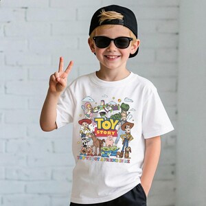 Op de afbeelding: Wit T-shirt met een kleurrijke afbeelding van Toy Story-personages, waaronder Woody en Buzz Lightyear. Het shirt heeft de tekst "TOY STORY" en "YOU'VE GOT A FRIEND IN ME". Het shirt wordt gedragen door een persoon die een zwarte pet en een zonnebril draagt.