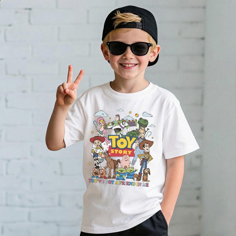 Op de afbeelding: Wit T-shirt met een kleurrijke afbeelding van Toy Story-personages, waaronder Woody en Buzz Lightyear. Het shirt heeft de tekst "TOY STORY" en "YOU'VE GOT A FRIEND IN ME". Het shirt wordt gedragen door een persoon die een zwarte pet en een zonnebril draagt.