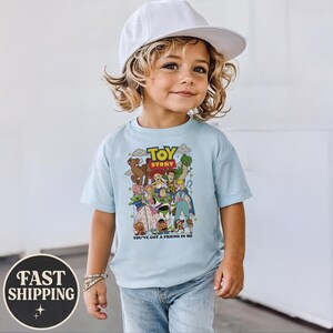 Puede incluir: Una camiseta azul claro con un estampado colorido de personajes de la pel&iacute;cula "Toy Story". La camiseta tiene las palabras "Toy Story" y "You've Got a Friend in Me" impresas. El ni&ntilde;o lleva una gorra de b&eacute;isbol blanca y vaqueros azules.