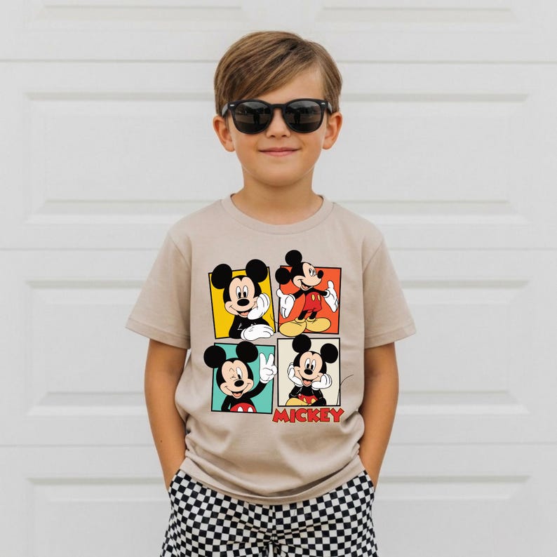 May include: A beige t-shirt with four panels depicting Mickey Mouse in various poses. The panels have yellow, red, and blue backgrounds. The word "Mickey" is printed in red. The child is wearing black sunglasses and checkered shorts.