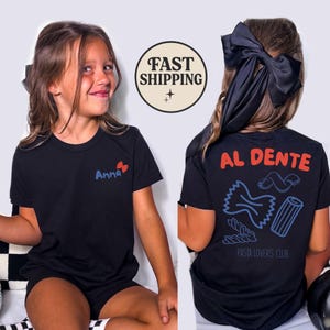May include: Black t-shirt with the name "Anna" in blue and a red heart design. The back of the shirt features the words "AL DENTE" in red, pasta illustrations, and "PASTA LOVERS CLUB" in blue. The image also includes a "FAST SHIPPING" graphic.