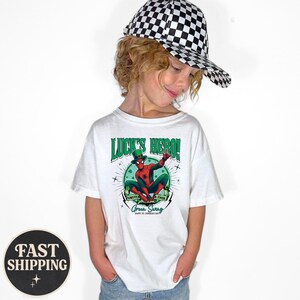 Puede incluir: Una camiseta blanca con un gr&aacute;fico de Spider-Man con un sombrero de duende verde. La camiseta tiene el texto "Luck's Hero!" y "Forever Green Swag" con un tema del D&iacute;a de San Patricio. El ni&ntilde;o lleva una gorra de b&eacute;isbol a cuadros blancos y negros.
