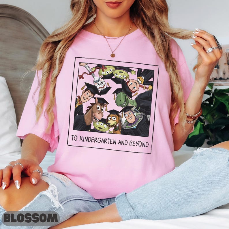 May include: A light pink t-shirt featuring a square graphic of Toy Story characters wearing graduation caps. The characters include Woody, Buzz Lightyear, Rex, Bullseye, and aliens. The text below the graphic reads TO KINDERGARTEN AND BEYOND.