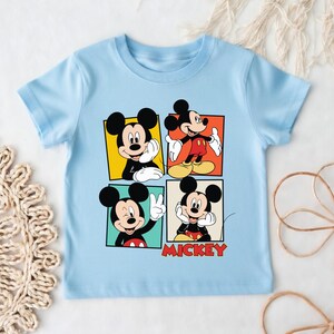 May include: Pale blue t-shirt with four panels showcasing Mickey Mouse in various poses. Each panel has a different coloured background: yellow, orange, green, and cream. The word "Mickey" is printed in red at the bottom.