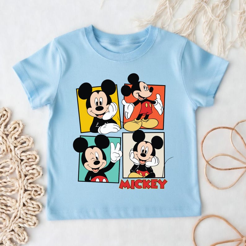May include: Pale blue t-shirt with four panels showcasing Mickey Mouse in various poses. Each panel has a different coloured background: yellow, orange, green, and cream. The word "Mickey" is printed in red at the bottom.