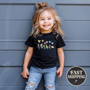 K&ouml;nnte beinhalten: Ein l&auml;chelndes Kind tr&auml;gt ein schwarzes T-Shirt mit einer bunten Grafik von Zeichentrickfiguren. Das Shirt zeigt eine Reihe von Figuren und Mickey Mouse-f&ouml;rmige Ballons. Das Kind tr&auml;gt eine blaue Jeans und steht vor einer grauen Wand.