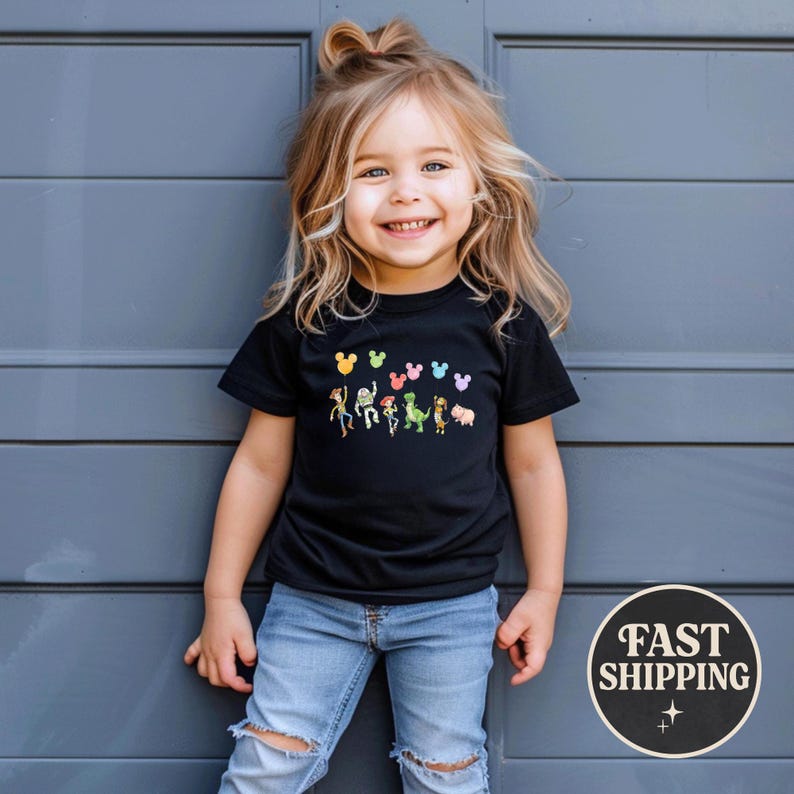 K&ouml;nnte beinhalten: Ein l&auml;chelndes Kind tr&auml;gt ein schwarzes T-Shirt mit einer bunten Grafik von Zeichentrickfiguren. Das Shirt zeigt eine Reihe von Figuren und Mickey Mouse-f&ouml;rmige Ballons. Das Kind tr&auml;gt eine blaue Jeans und steht vor einer grauen Wand.