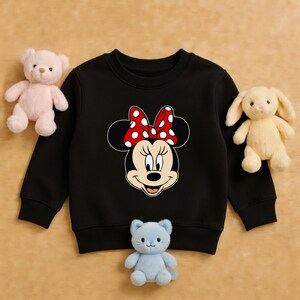Może przedstawiać: Czarna bluza dziecięca z grafiką Myszki Minnie. Grafika przedstawia twarz Myszki Minnie z czerwoną kokardą w białe grochy. Bluza otoczona jest pluszakami, w tym r&oacute;żowym misiem, ż&oacute;łtym kr&oacute;likiem i niebieskim kotem.