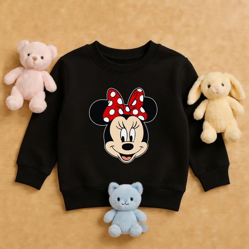 Może przedstawiać: Czarna bluza dziecięca z grafiką Myszki Minnie. Grafika przedstawia twarz Myszki Minnie z czerwoną kokardą w białe grochy. Bluza otoczona jest pluszakami, w tym r&oacute;żowym misiem, ż&oacute;łtym kr&oacute;likiem i niebieskim kotem.