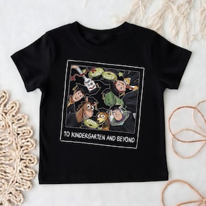 May include: A black short-sleeved t-shirt featuring a square graphic print of various animated characters wearing graduation caps. The text "TO KINDERGARTEN AND BEYOND" is printed in white capital letters at the bottom of the square frame.