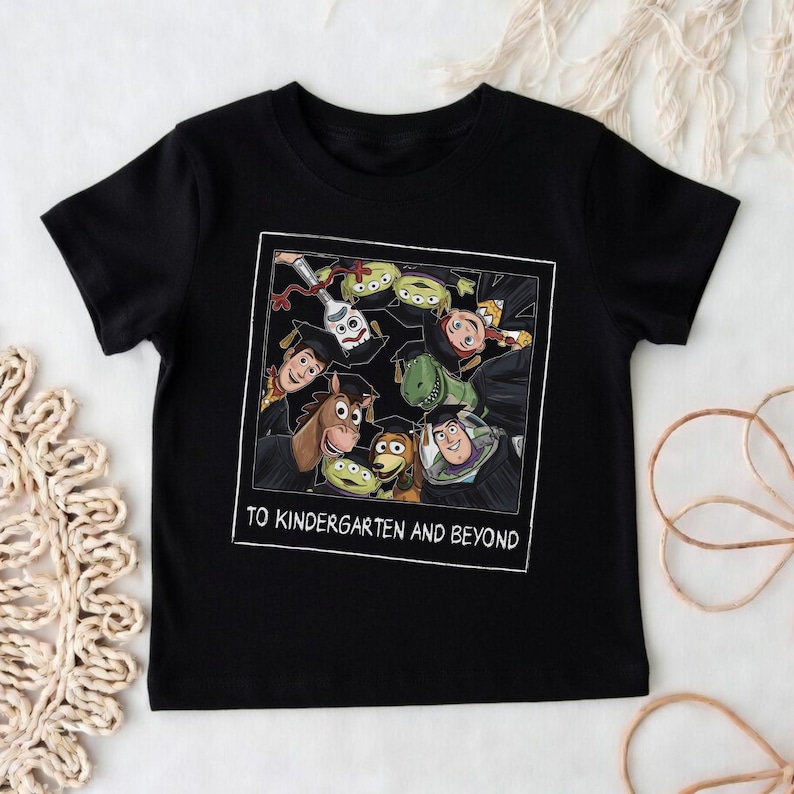 May include: A black short-sleeved t-shirt featuring a square graphic print of various animated characters wearing graduation caps. The text "TO KINDERGARTEN AND BEYOND" is printed in white capital letters at the bottom of the square frame.