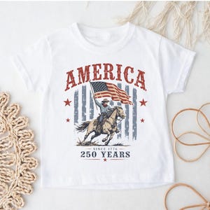 May include: A white t-shirt featuring a graphic print of a cowboy riding a horse while holding an American flag. The text reads AMERICA at the top and SINCE 1776 250 YEARS at the bottom, accented by red stars and a faded blue striped background.