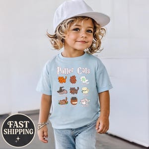 May include: A light blue t-shirt with the text "Potter Cats" and illustrations of cartoon cats. The shirt features a variety of cat designs, each with a unique color and style. The shirt is worn by a child.
