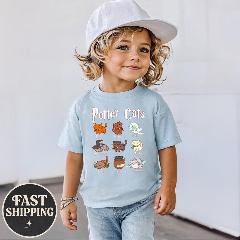 May include: A light blue t-shirt with the text "Potter Cats" and illustrations of cartoon cats. The shirt features a variety of cat designs, each with a unique color and style. The shirt is worn by a child.