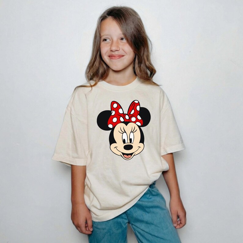 Może przedstawiać: Kremowa koszulka z grafiką przedstawiającą twarz Myszki Minnie. Kresk&oacute;wkowa postać ma czarne uszy, czerwoną kokardę w białe grochy i uśmiechniętą minę. Koszulka jest noszona przez osobę w niebieskich dżinsach.