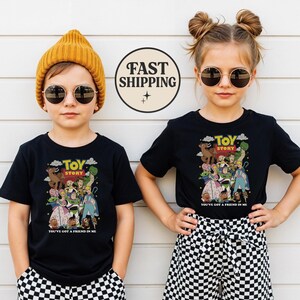 Puede incluir: Dos ni&ntilde;os con camisetas negras con un gr&aacute;fico colorido de Toy Story. Las camisetas presentan el t&iacute;tulo de la pel&iacute;cula y la frase "You've Got a Friend in Me". Uno lleva un gorro amarillo, el otro tiene el pelo recogido en mo&ntilde;os, y ambos llevan gafas de sol.