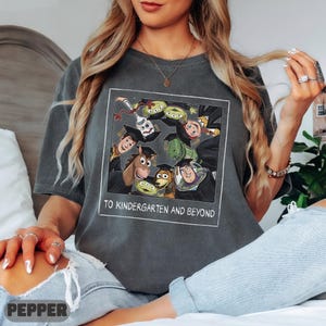 May include: A charcoal gray short-sleeve t-shirt featuring a square graphic of Toy Story characters wearing graduation caps. The text below the characters reads TO KINDERGARTEN AND BEYOND. The shirt has a crew neckline and a relaxed, vintage-style fit.