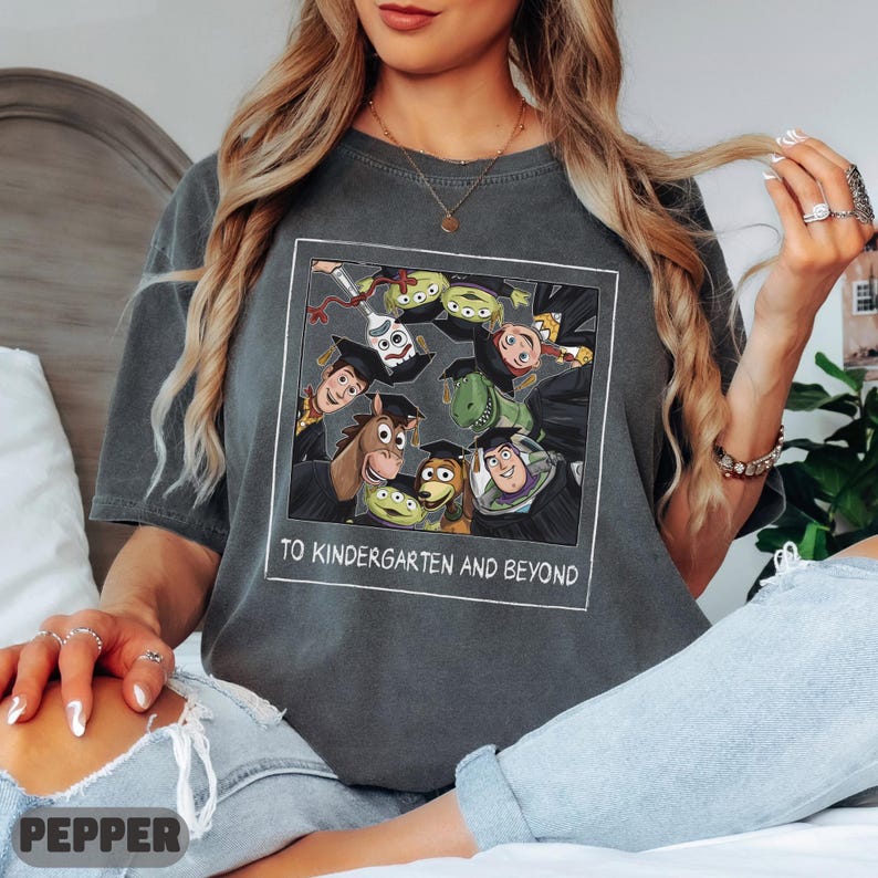 May include: A charcoal gray short-sleeve t-shirt featuring a square graphic of Toy Story characters wearing graduation caps. The text below the characters reads TO KINDERGARTEN AND BEYOND. The shirt has a crew neckline and a relaxed, vintage-style fit.