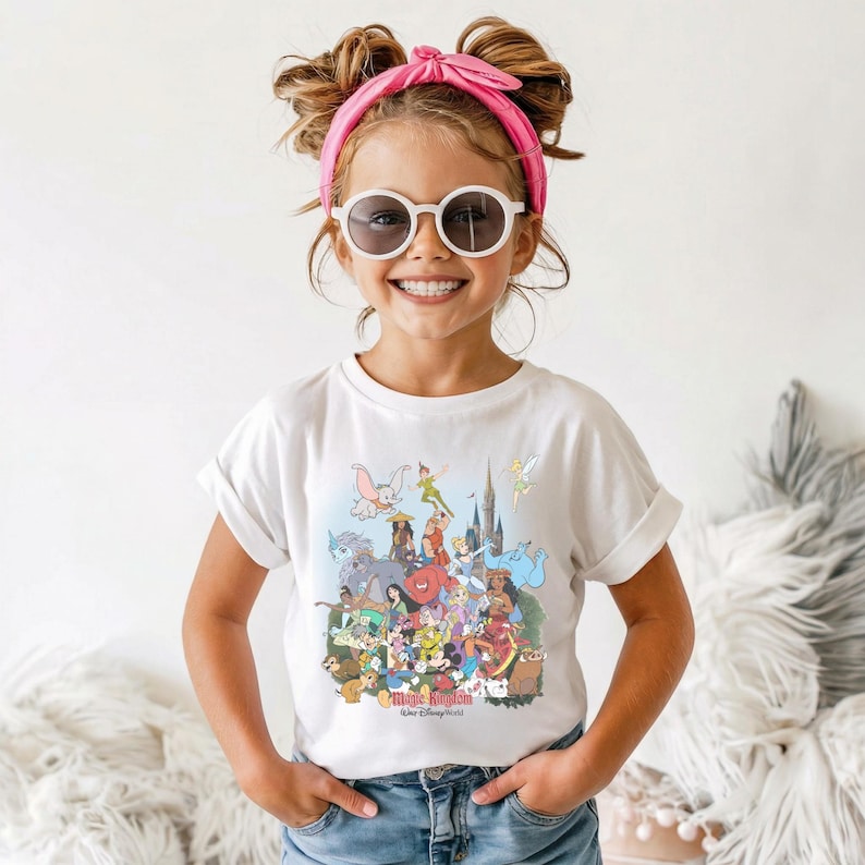 May include: A white t-shirt featuring a colorful graphic of various Disney characters, including Mickey Mouse, Dumbo, and Peter Pan. The shirt has rolled-up sleeves and is worn by a young person. The text "Magic Kingdom Walt Disney World" is visible.