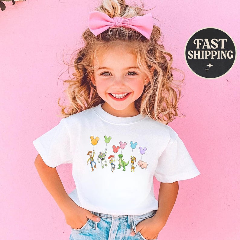 K&ouml;nnte beinhalten: Ein wei&szlig;es T-Shirt mit einer Cartoon-Grafik von Figuren, die Mickey Mouse-f&ouml;rmige Ballons halten. Das Design umfasst Charaktere wie Woody und Buzz Lightyear. Das T-Shirt wird von einem kleinen Kind mit einer rosa Schleife im Haar getragen.