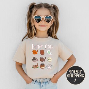May include: A beige t-shirt featuring the text "Potter Cats" above cartoon illustrations of cats in various poses. The shirt has a round neck and short sleeves. The girl is wearing heart-shaped sunglasses.