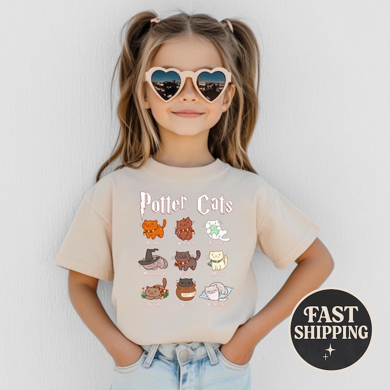 May include: A beige t-shirt featuring the text "Potter Cats" above cartoon illustrations of cats in various poses. The shirt has a round neck and short sleeves. The girl is wearing heart-shaped sunglasses.