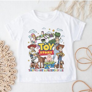Op de afbeelding: Wit T-shirt met een kleurrijke afbeelding van personages uit de animatiefilm "Toy Story". Het ontwerp bevat Woody, Buzz Lightyear en andere personages, met de tekst "TOY STORY" en "YOU'VE GOT A FRIEND IN ME."