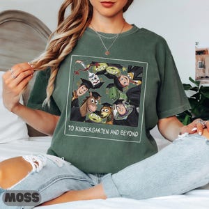May include: A moss green short-sleeve t-shirt featuring a graphic print of Toy Story characters wearing graduation caps. The design includes the text TO KINDERGARTEN AND BEYOND inside a white square border, showcasing Woody, Buzz Lightyear, and friends.