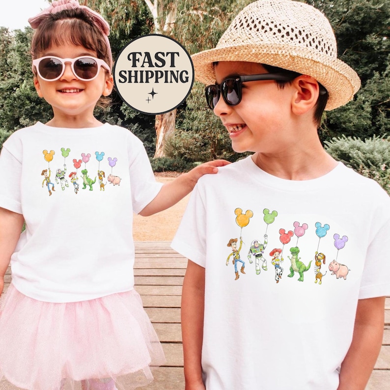 K&ouml;nnte beinhalten: Zwei Kinder tragen wei&szlig;e T-Shirts mit einer bunten Cartoon-Grafik von Figuren, die ballonf&ouml;rmige Ballons halten. Die Grafik zeigt einen Cowboy, einen Weltraumranger und einen Dinosaurier. Ein Kind tr&auml;gt einen rosa T&uuml;llrock und eine Sonnenbrille.