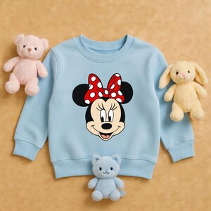 Może przedstawiać: Jasnoniebieska bluza z nadrukiem Myszki Minnie, z czerwoną kokardą w białe grochy. Na zdjęciu znajduje się r&oacute;wnież r&oacute;żowy miś, ż&oacute;łty kr&oacute;lik i niebieska pluszowa zabawka w kształcie kota.